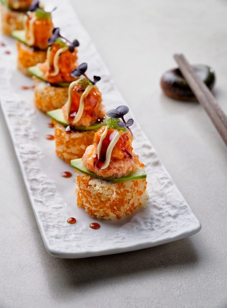 Elegant studio food photography by a food photographer in Dubai featuring a premium maki sushi roll presentation. A row of five identical sushi pieces is arranged in a precise diagonal line across a textured, long white ceramic platter. Each piece features a crispy tempura-flaked and orange tobiko (fish roe) coated base, topped with a thin slice of fresh green avocado and a mound of spicy minced salmon or tuna tartar. The sushi is meticulously garnished with a drizzle of white kewpie mayonnaise, a glossy dark unagi sauce drop, green caviar beads, and a single purple micro-herb leaf. In the blurred background, a pair of wooden chopsticks rests on a dark ceramic holder on a neutral grey concrete or stone tabletop under soft, directional top-left studio lighting.