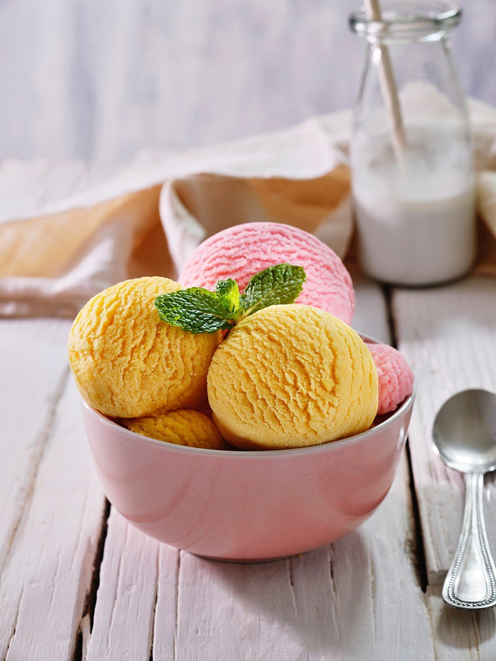 A premium commercial food photograph by Orkun Orcan featuring perfectly sculpted scoops of mango and strawberry ice cream in a pastel pink bowl, garnished with fresh mint on a rustic white wooden table.